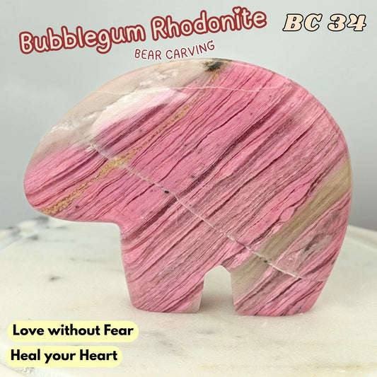 A carved pink rhodonite bear figurine with natural striated patterns, displayed on a light marble surface with text overlays reading 'Bubblegum Rhodonite', 'Bear Carving', 'BC 34', and 'Love without Fear Heal your Heart'.