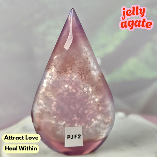 A teardrop-shaped jelly agate crystal with a pinkish-purple hue and glittery internal inclusions, displayed on a soft white surface with text overlays reading 'jelly agate', 'Attract Love', and 'Heal Within'.