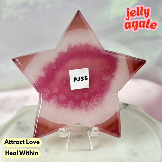 A star-shaped jelly agate crystal on a clear acrylic stand, featuring a pink and magenta geode pattern with a white label reading 'PJS5'. The background is a soft-focus light gray and white surface with decorative text overlays 'jelly agate' and 'Attract Love Heal Within'.