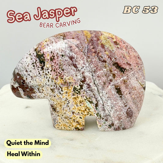 A carved sea jasper bear figurine with natural multicolored stone patterns in pink, gold, white, and brown, displayed on a light marble surface against a neutral gray background. Text overlays include 'Sea Jasper BEAR CARVING', 'BC 53', and 'Quiet the Mind Heal Within'.