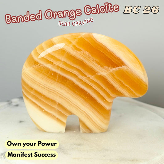 A carved orange-yellow stone bear figurine with banded calcite-like striations, shown on a light marble surface against a gray background. Text overlays read 'Banded Orange Calcite', 'BC 26', 'BEAR CARVING', and 'Own your Power Manifest Success'.