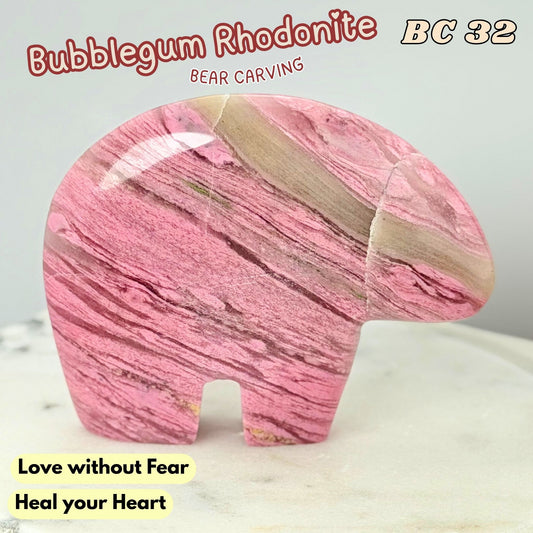 A polished pink rhodonite carved bear figurine with natural banding, displayed on a light marble surface against a neutral light gray background. Text overlays include 'Bubblegum Rhodonite', 'BC 32', 'Bear Carving', and 'Love without Fear Heal your Heart'.