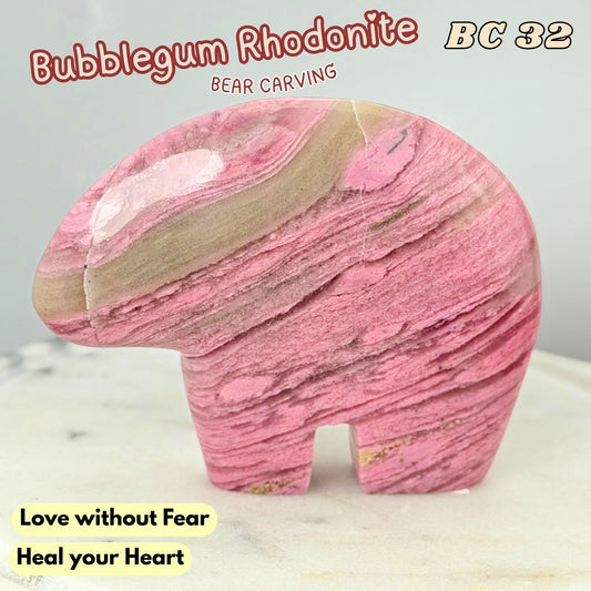 A carved pink rhodonite bear figurine with natural stone banding, displayed on a light marble surface against a pale gray background. Text overlays identify the stone as Bubblegum Rhodonite and include the carving number BC 32 and the phrase 'Love without Fear Heal your Heart'.