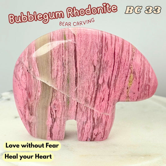 A carved pink rhodonite bear figurine with natural stone veining, displayed on a light marble surface against a light gray background. Text overlays include 'Bubblegum Rhodonite', 'BC 33', 'Love without Fear', and 'Heal your Heart'.
