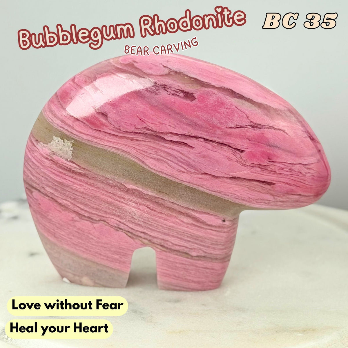 A polished pink rhodonite carved bear figurine with natural banding, displayed on a light surface against a pale background. Text overlays include 'Bubblegum Rhodonite', 'BC 35', 'Love without Fear', and 'Heal your Heart'.