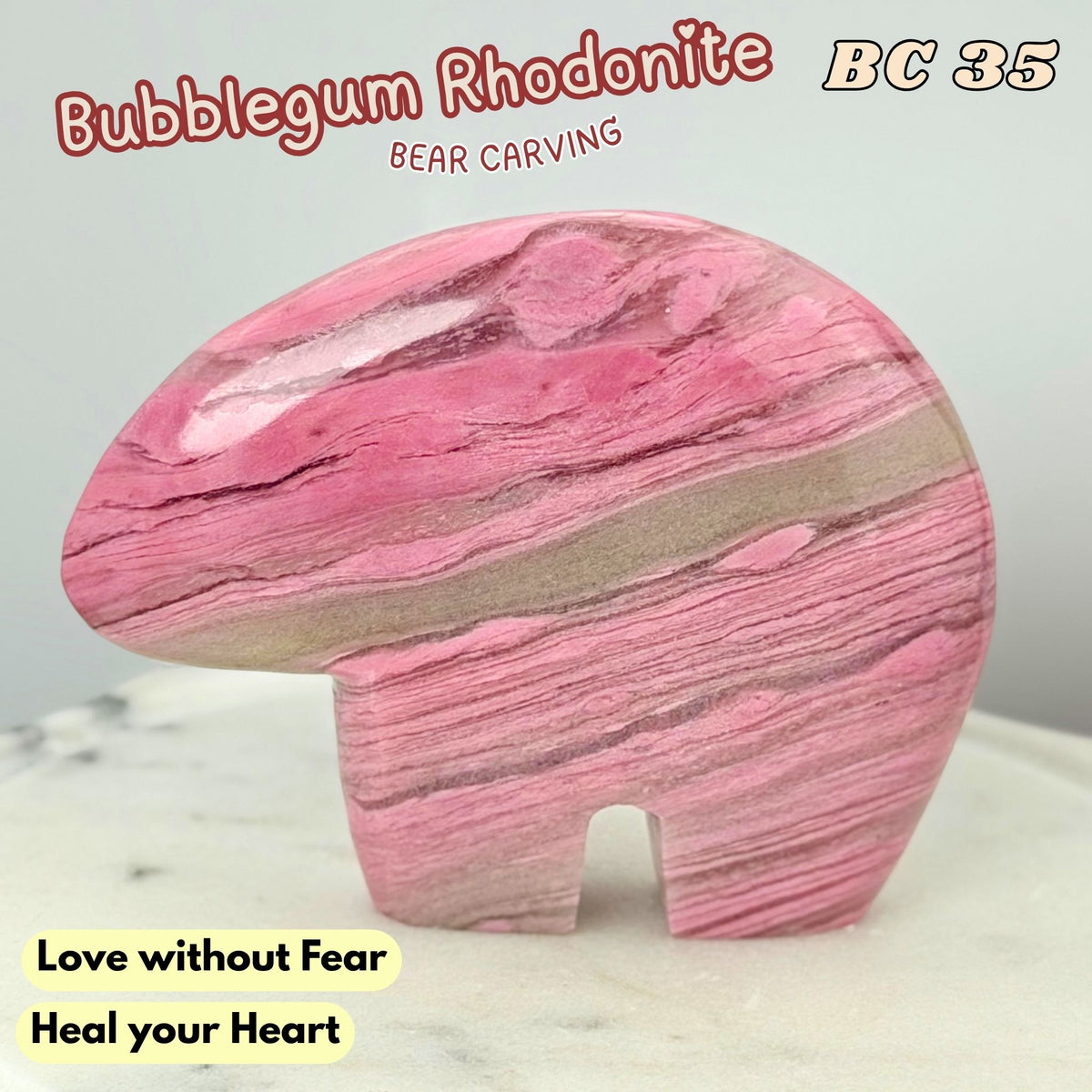 A polished pink rhodonite bear carving with natural marbled bands, displayed on a light marble surface against a pale gray background. Text overlays read 'Bubblegum Rhodonite', 'Bear Carving', 'BC 35', and 'Love without Fear Heal your Heart'.