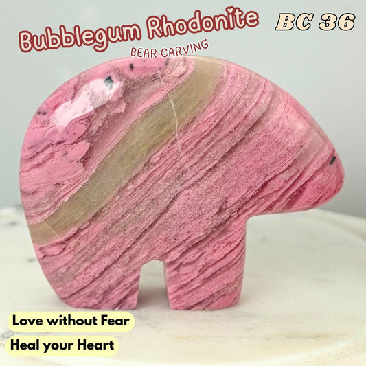 A carved bear-shaped stone ornament made of pink and beige bubblegum rhodonite, displayed on a light marble surface with text overlays.