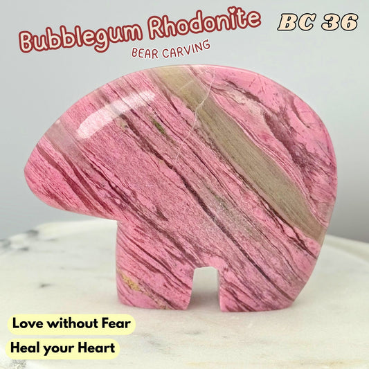 A polished pink rhodonite carved bear figurine with natural marbled veining, displayed on a light marble surface against a neutral gray background. Text overlays read 'Bubblegum Rhodonite', 'BC 36', 'BEAR CARVING', and 'Love without Fear Heal your Heart'.