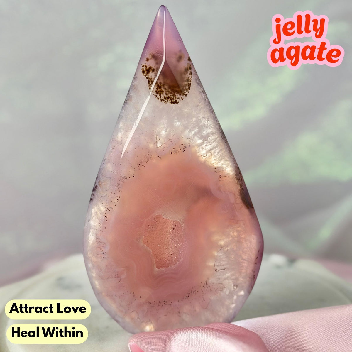 A teardrop-shaped jelly agate crystal with pink and purple agate bands and a gold vein, held by a hand against a soft iridescent background. Text overlays read 'jelly agate', 'Attract Love', and 'Heal Within'.