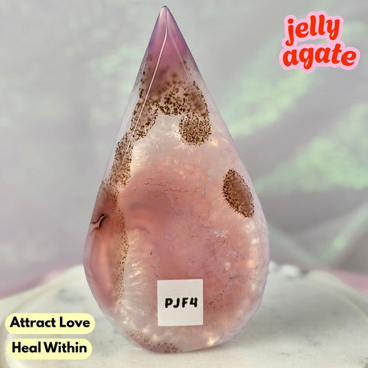 A teardrop-shaped polished jelly agate crystal with pink and purple tones and brown inclusions, displayed on a light surface with text overlays reading 'jelly agate', 'Attract Love', 'Heal Within', and a label 'PJP4'.