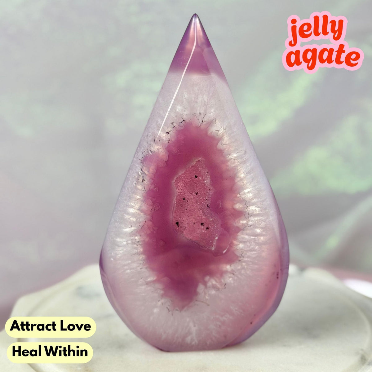 A teardrop-shaped jelly agate crystal with a pink and purple geode-like pattern, displayed on a white surface with text overlays reading 'jelly agate', 'Attract Love', and 'Heal Within'.