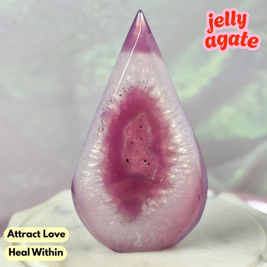 A teardrop-shaped jelly agate crystal with a pink and purple geode-like pattern, displayed on a white surface with text overlays reading 'jelly agate', 'Attract Love', and 'Heal Within'.