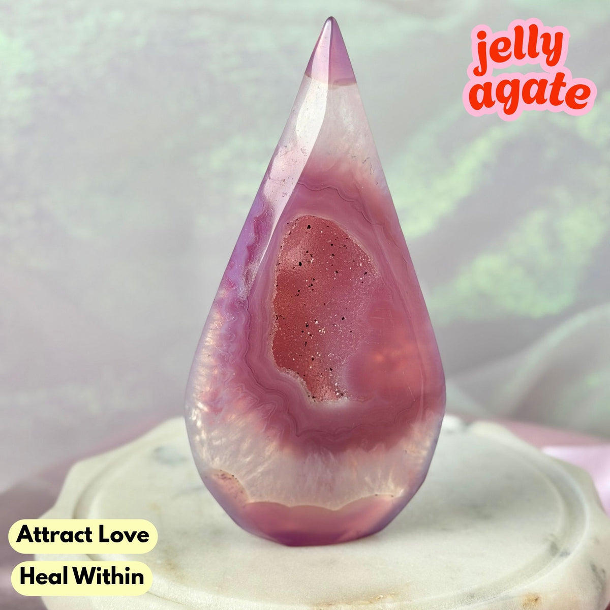 A teardrop-shaped polished jelly agate crystal point with pink, purple, and white banded patterns, displayed on a white marble pedestal. Text overlays read 'jelly agate' and 'Attract Love Heal Within'.