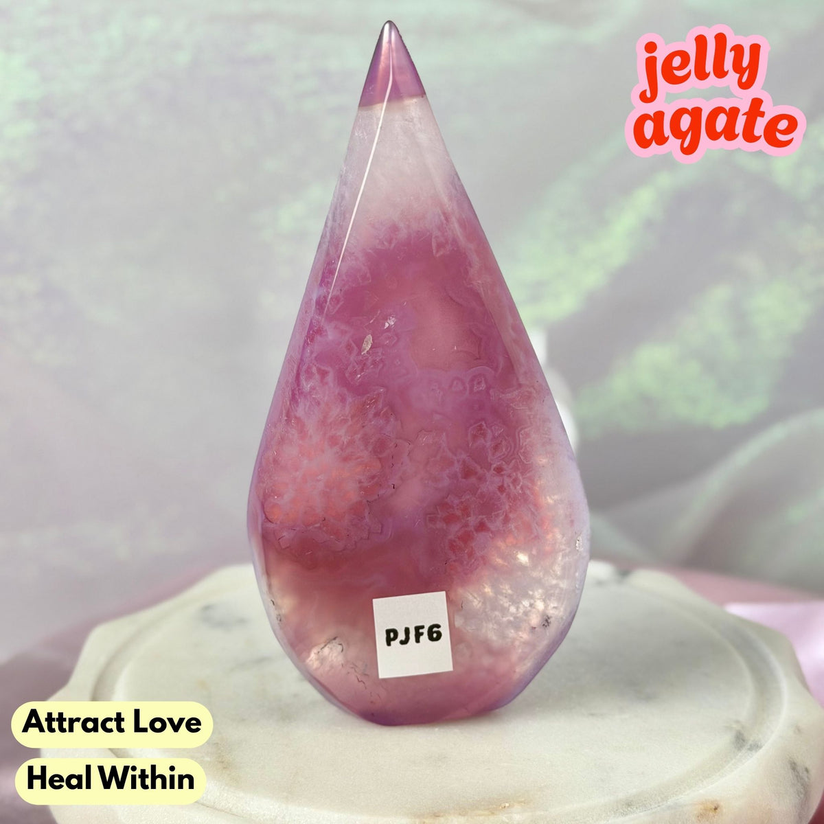 A teardrop-shaped polished jelly agate crystal tower with pink and purple marbled patterns, displayed on a white stone pedestal. Text overlays read 'jelly agate', 'Attract Love', and 'Heal Within'.
