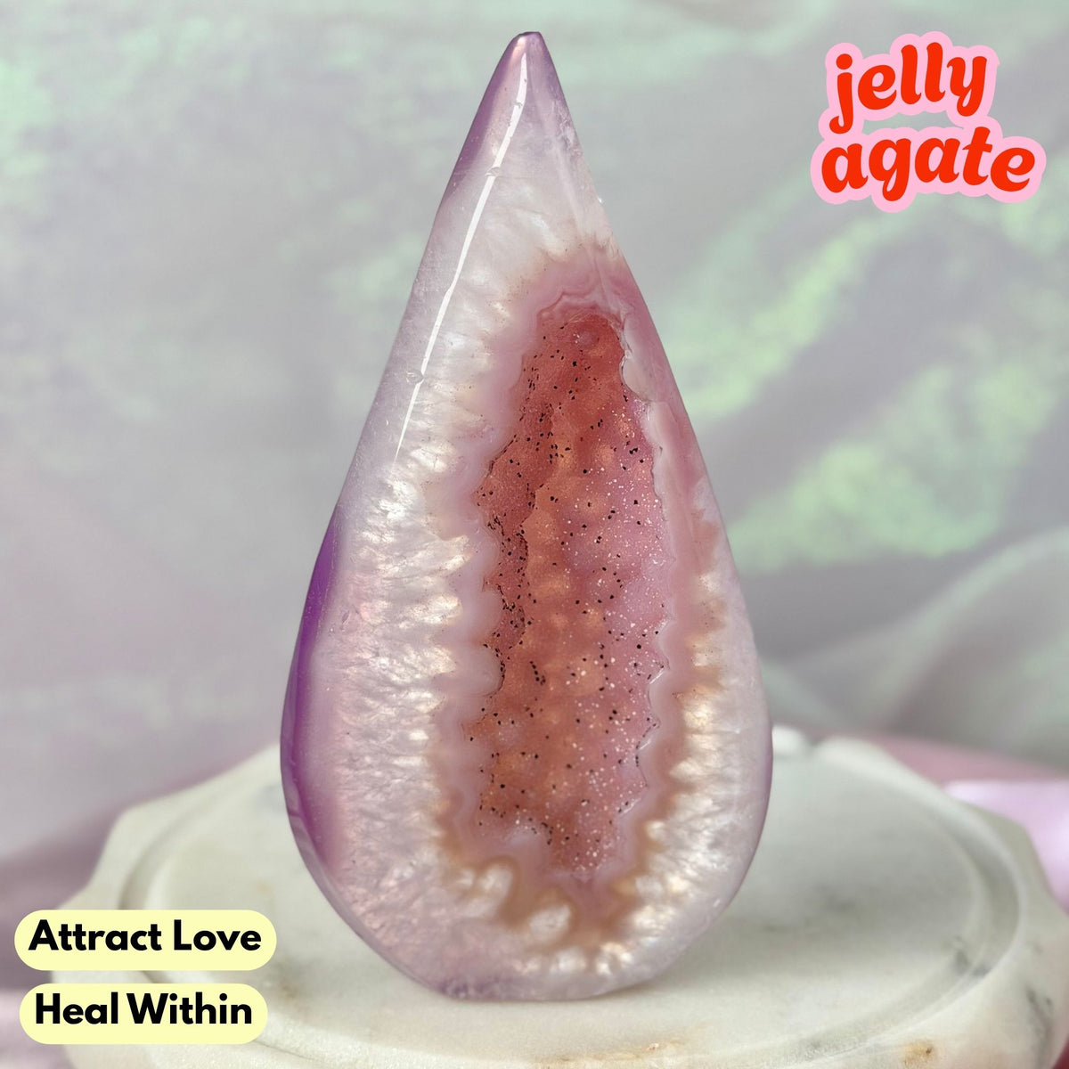 A polished teardrop-shaped jelly agate crystal with a purple outer layer and a pinkish-brown geode-like center, displayed on a white pedestal. Text overlays read 'jelly agate', 'Attract Love', and 'Heal Within'.