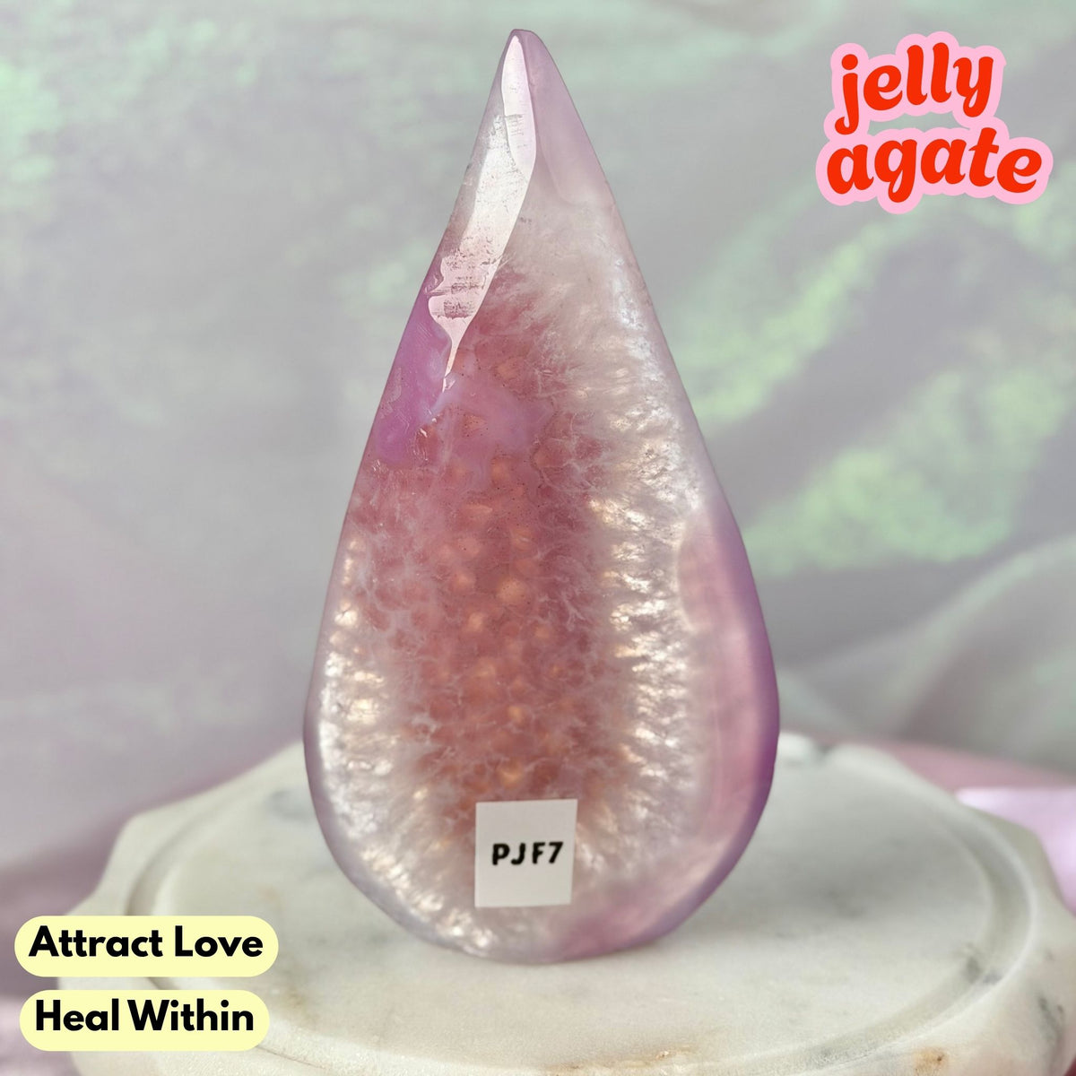 A teardrop-shaped polished jelly agate crystal with a pinkish-purple outer layer and a translucent inner cavity filled with small orange-brown spheres, displayed on a white pedestal. Text overlays read 'jelly agate', 'Attract Love Heal Within', and 'PJJ7'.