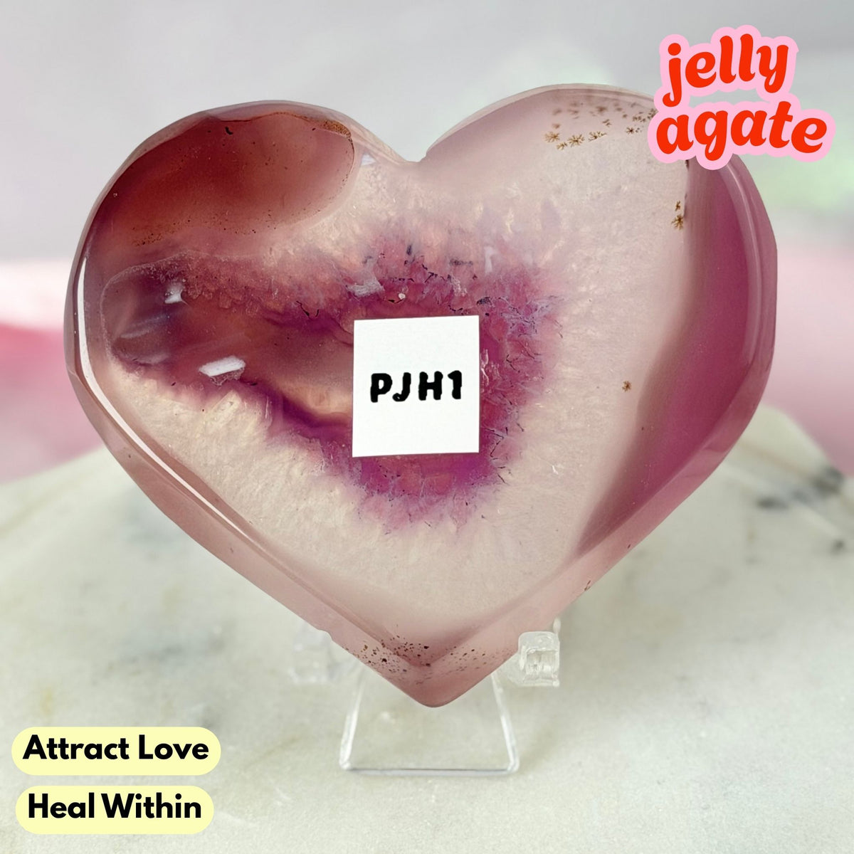 A heart-shaped polished jelly agate crystal on a clear acrylic stand, featuring pink and purple agate bands with tiny gold inclusions. A white label with 'PJM1' is placed on the center, with text overlays 'jelly agate' and 'Attract Love Heal Within'.