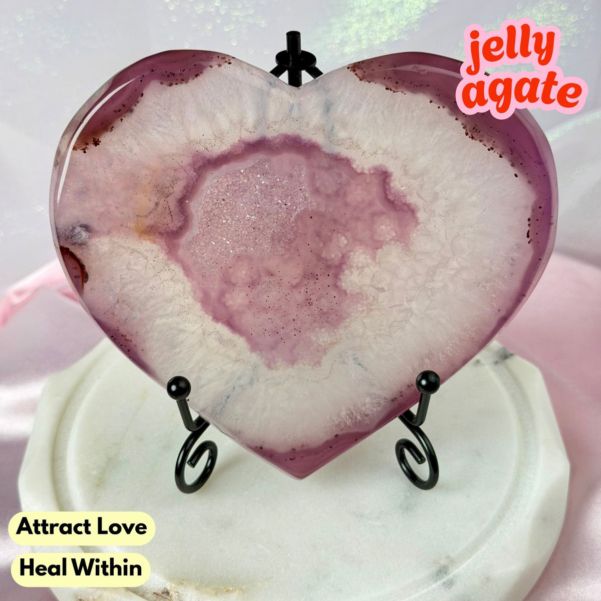 Heart-shaped polished agate stone display on a small black stand, with a pink and white marbled center and purple outer edges. The stone is shown against a soft pastel background with text overlays reading 'jelly agate' and 'Attract Love Heal Within'.