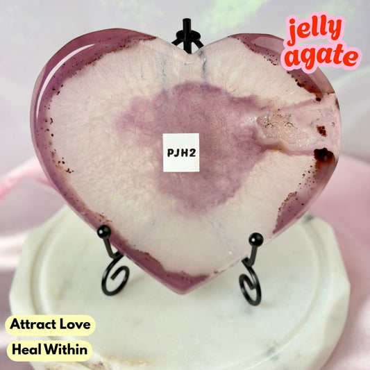 Heart-shaped polished jelly agate crystal on a small black stand, displayed on a white marble base. The crystal features a pinkish-purple outer ring and a pale pink to off-white center with natural marbling.