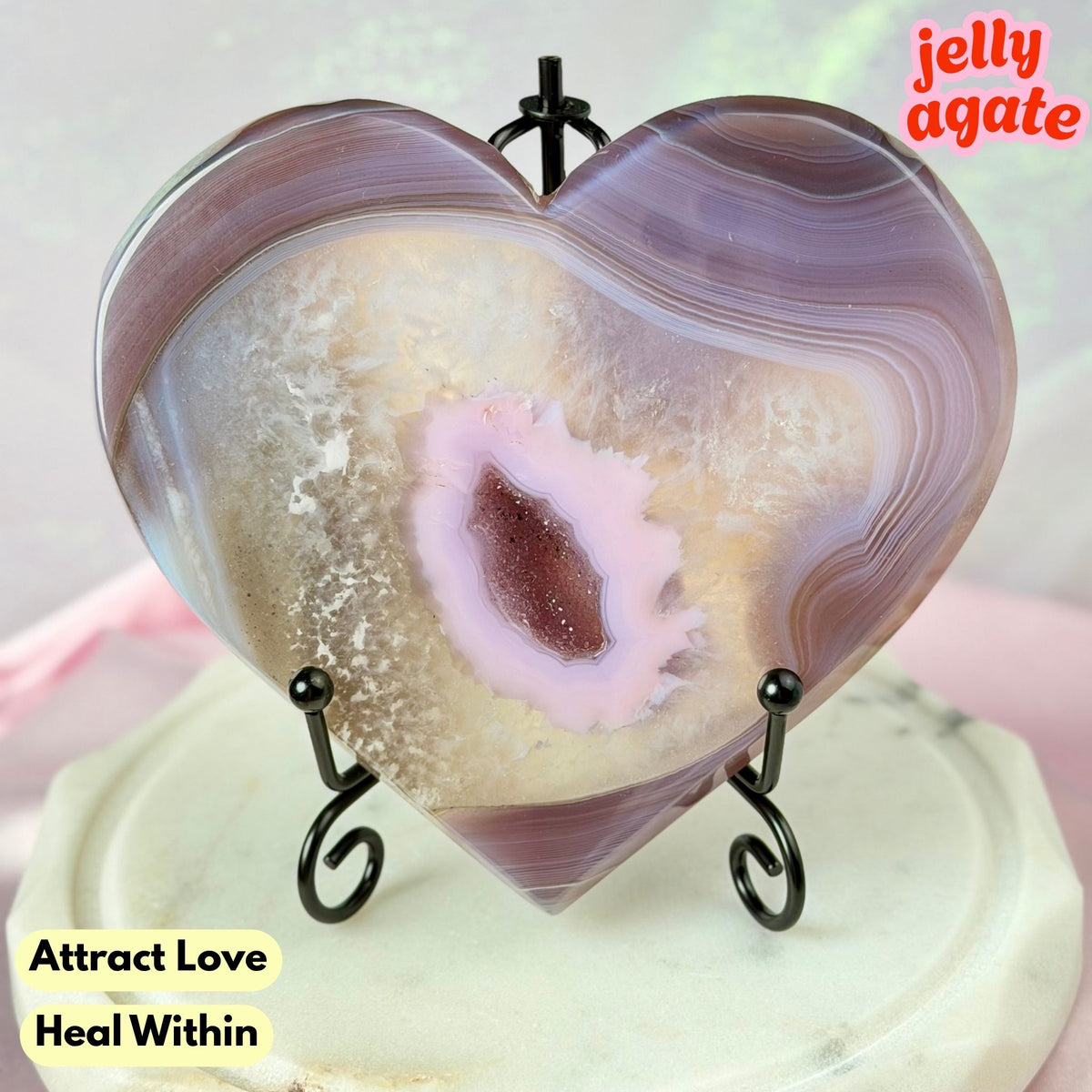 Heart-shaped jelly agate crystal on a small black metal stand, featuring purple and beige banded agate patterns with a pale pink center. The crystal is displayed on a light-colored surface with text overlays reading 'jelly agate' and 'Attract Love Heal Within'.