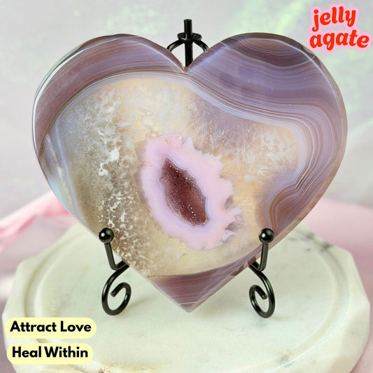 Heart-shaped jelly agate crystal on a small black metal stand, featuring purple and beige banded agate patterns with a pale pink center. The crystal is displayed on a light-colored surface with text overlays reading 'jelly agate' and 'Attract Love Heal Within'.