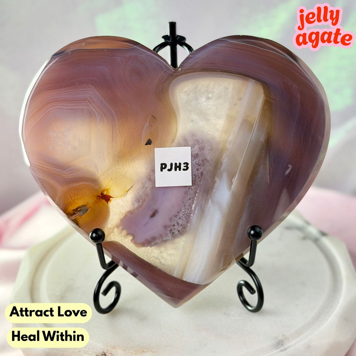 Heart-shaped polished agate crystal on a black metal stand, with a small white label reading 'PJM3' and text overlays 'Attract Love Heal Within' and 'jelly agate'.