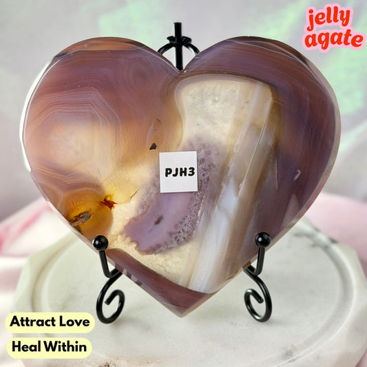 Heart-shaped polished agate crystal on a black metal stand, with a small white label reading 'PJM3' and text overlays 'Attract Love Heal Within' and 'jelly agate'.