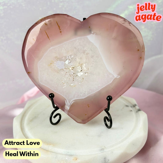 Heart-shaped pink agate crystal display piece on a small black wire stand, placed on a round white marble base. The agate has a smooth polished surface with a white crystalline center and tiny gold flecks.