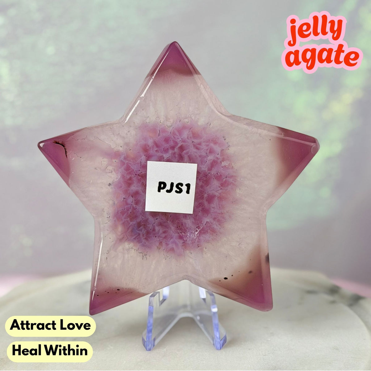 A star-shaped jelly agate crystal on a clear acrylic stand, featuring a pink and purple marbled pattern. The crystal has a white label in the center that reads 'PJS1' and is set against a soft, blurry greenish background with text overlays 'jelly agate' and 'Attract Love Heal Within'.
