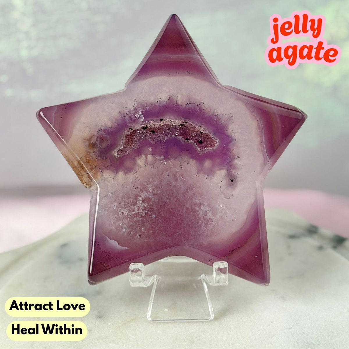 A star-shaped jelly agate crystal on a clear acrylic display stand, featuring a purple and pink agate geode pattern with a central brown band. The background is a soft-focus light green and pink gradient, with text overlays reading 'jelly agate' and 'Attract Love Heal Within'.
