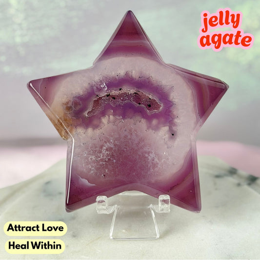 A star-shaped jelly agate crystal on a clear acrylic display stand, featuring a purple and pink agate geode pattern with a central brown band. The background is a soft-focus light green and pink gradient, with text overlays reading 'jelly agate' and 'Attract Love Heal Within'.