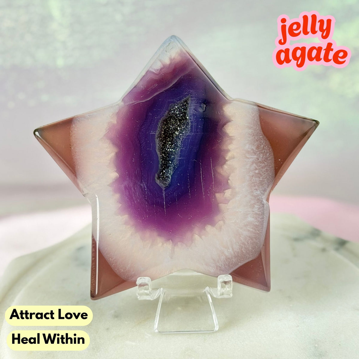 A star-shaped polished jelly agate crystal on a clear acrylic stand, featuring a purple and white geode pattern with a black glittery center. The background is a soft, blurry pastel pink and green, with text overlays reading 'jelly agate', 'Attract Love', and 'Heal Within'.