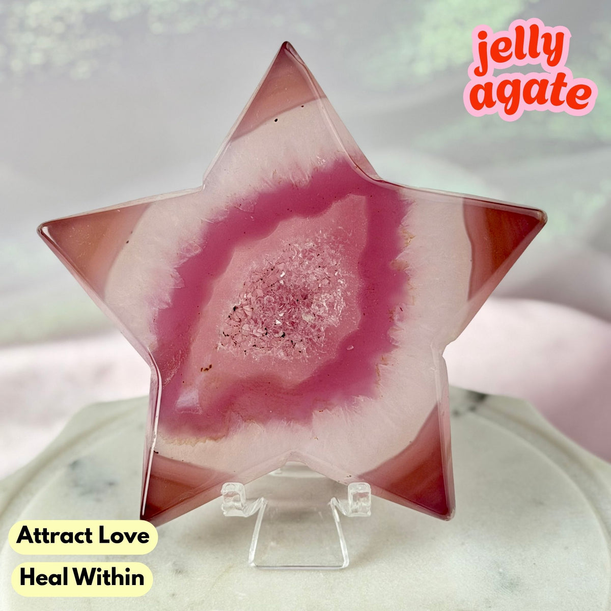 A star-shaped jelly agate crystal display on a clear acrylic stand, featuring a pink and white agate pattern with a glittery pink center. The background is a soft, blurred light gray and white surface with decorative text overlays reading 'jelly agate', 'Attract Love Heal Within', and 'Love'.