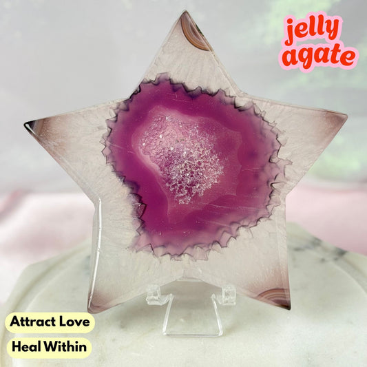 A star-shaped jelly agate crystal display piece on a clear acrylic stand, featuring a vibrant magenta-purple center with white crystalline inclusions and a translucent pale pink outer edge. Text overlays read 'jelly agate', 'Attract Love', and 'Heal Within'.