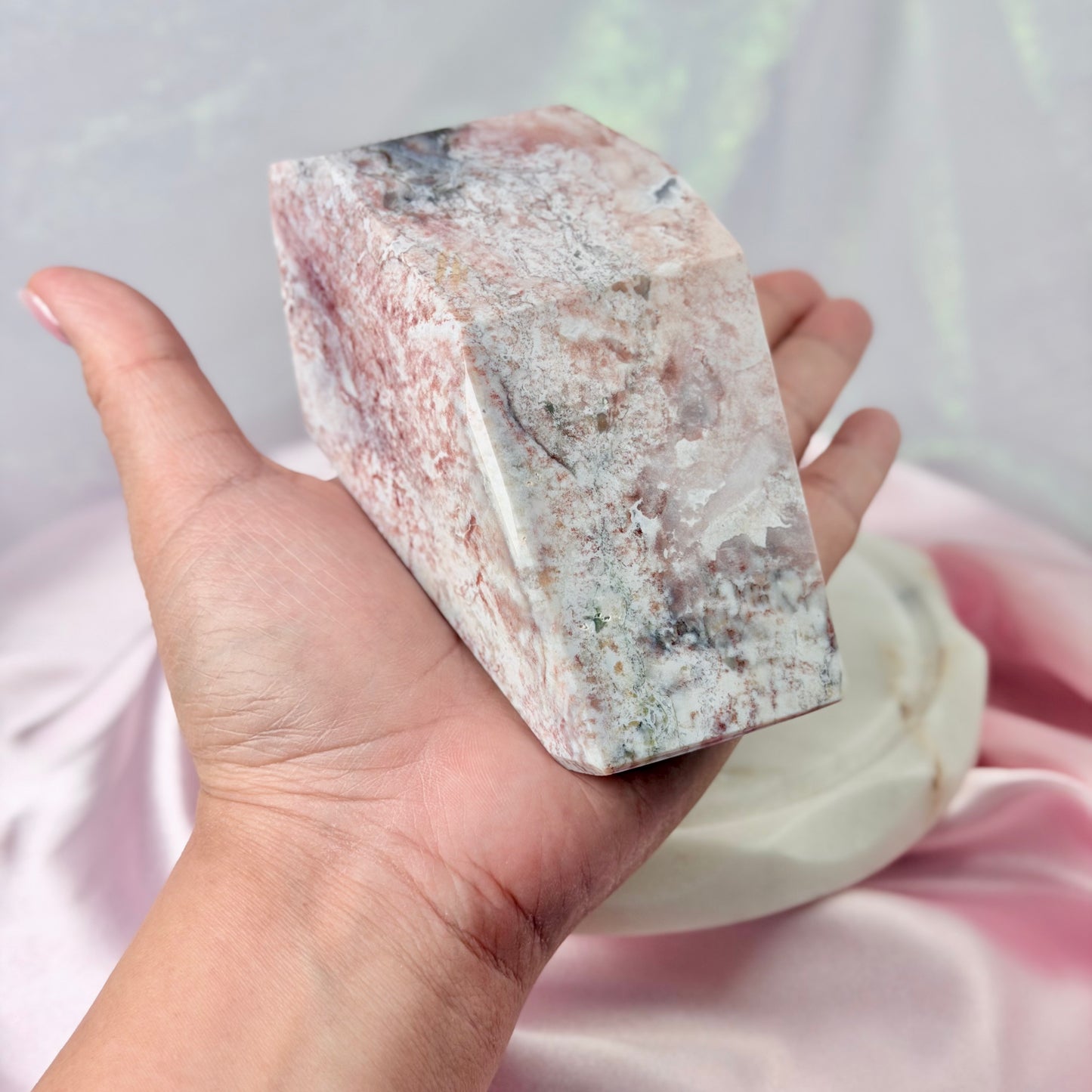 A hand holding a polished pinkish-white crystal tower with natural mineral textures, set against a soft pink draped fabric background.