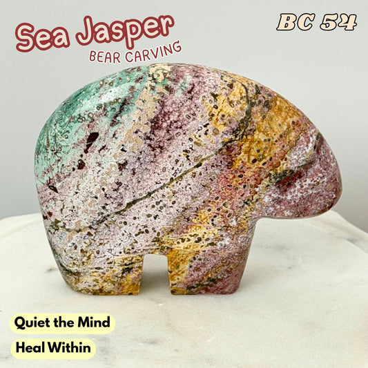 A carved sea jasper bear figurine with a marbled pattern of green, pink, yellow, and white, displayed on a light marble surface against a neutral gray background. Text overlays include 'Sea Jasper BEAR CARVING', 'BC 54', and 'Quiet the Mind Heal Within'.